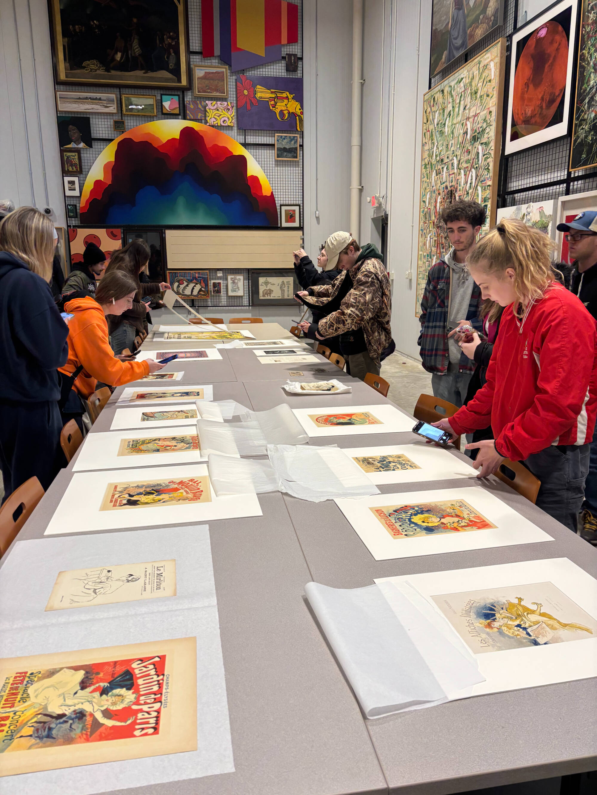 A group of college students in an art storage facility. They are looking at original French prints from the end of the 19th century. Some of them are taking photos with their phones.